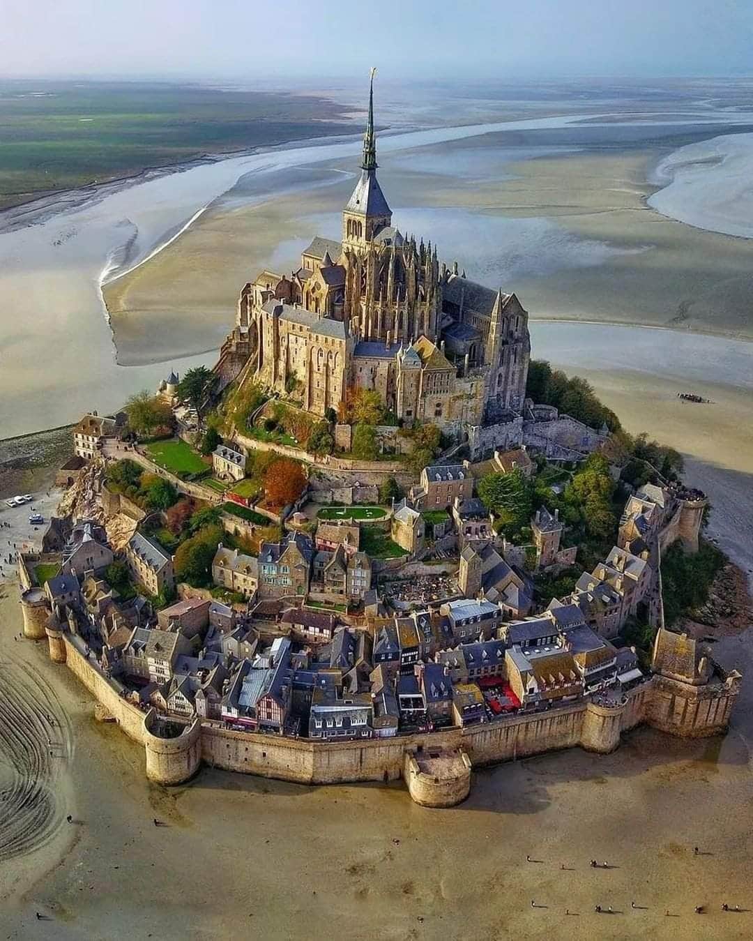 An aerial view showcases a historic island with a towering abbey at its peak, surrounded by medieval buildings and fortified walls. The landscape features sandy shores and water channels, highlighting the island's unique location. Green trees add a touch of color to the scene.