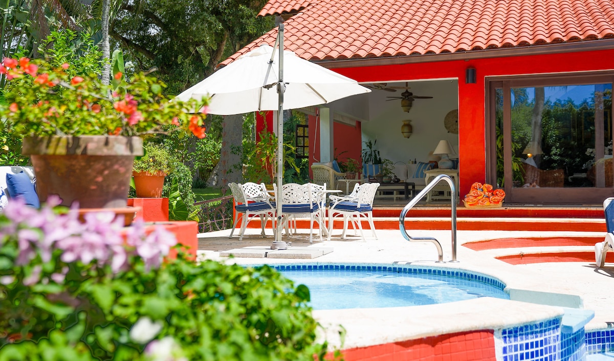 The image showcases a vibrant outdoor area featuring a hot tub beside a spacious patio. Surrounding lush greenery and colorful flowers enhance the scene. A large umbrella provides shade over a dining set, with a red-roofed villa visible in the background.