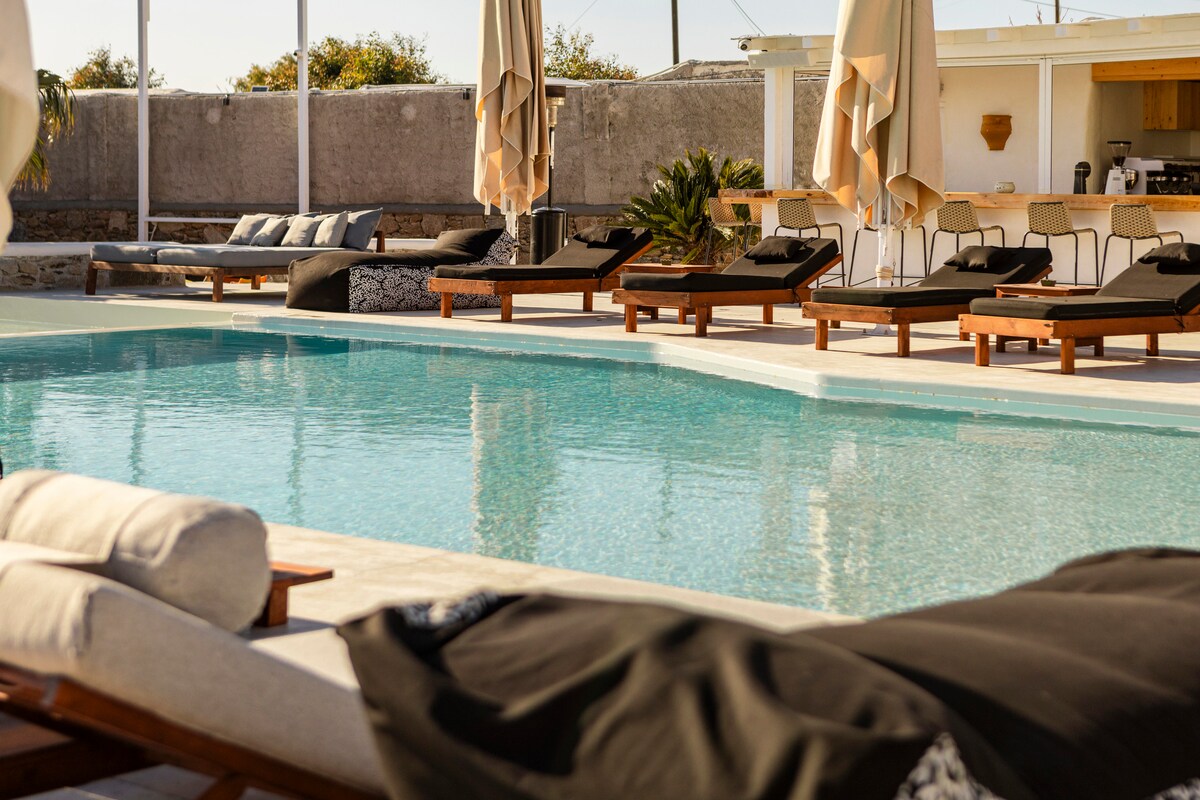 A serene pool area is presented, featuring a glistening water surface reflecting the sunlight. Multiple lounge chairs are arranged around the pool, accompanied by shaded areas from large umbrellas. A bar seating area is visible in the background, surrounded by lush greenery.