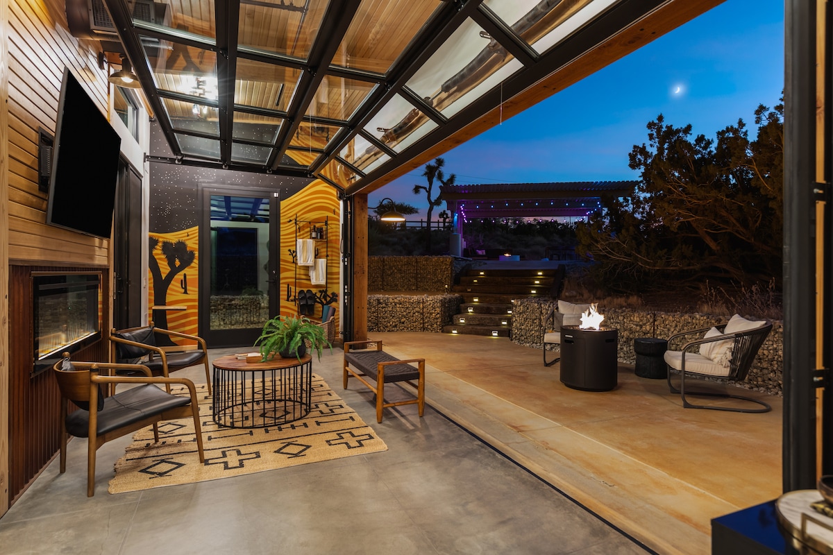 An open sunroom features a retractable glass wall, allowing easy access to the outdoors. Comfortable seating arrangements include two armchairs and a coffee table on a patterned rug. Soft lighting enhances the serene atmosphere, with a fire pit visible outside surrounded by desert landscaping.