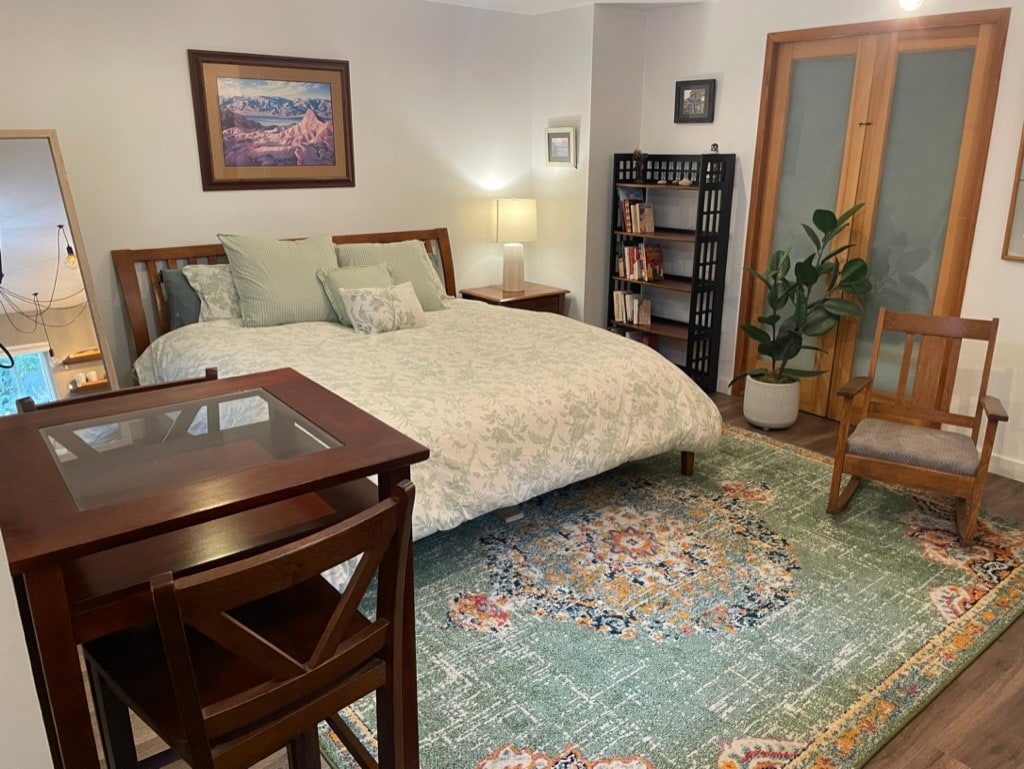 The guest unit features a spacious bed adorned with patterned bedding, complemented by soft pillows. A cozy area rug adds warmth underfoot. A wooden chair and table create a small dining space, while a bookshelf hosts a selection of books. Natural light enhances the welcoming environment.