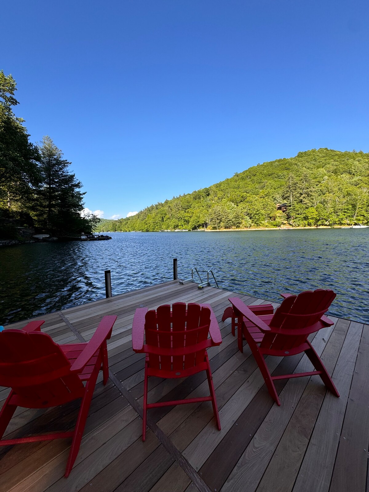 A wooden dock extends over the tranquil lake, featuring three bright red Adirondack chairs facing the water. Lush green hills can be seen in the background under a clear blue sky, creating a peaceful natural setting.