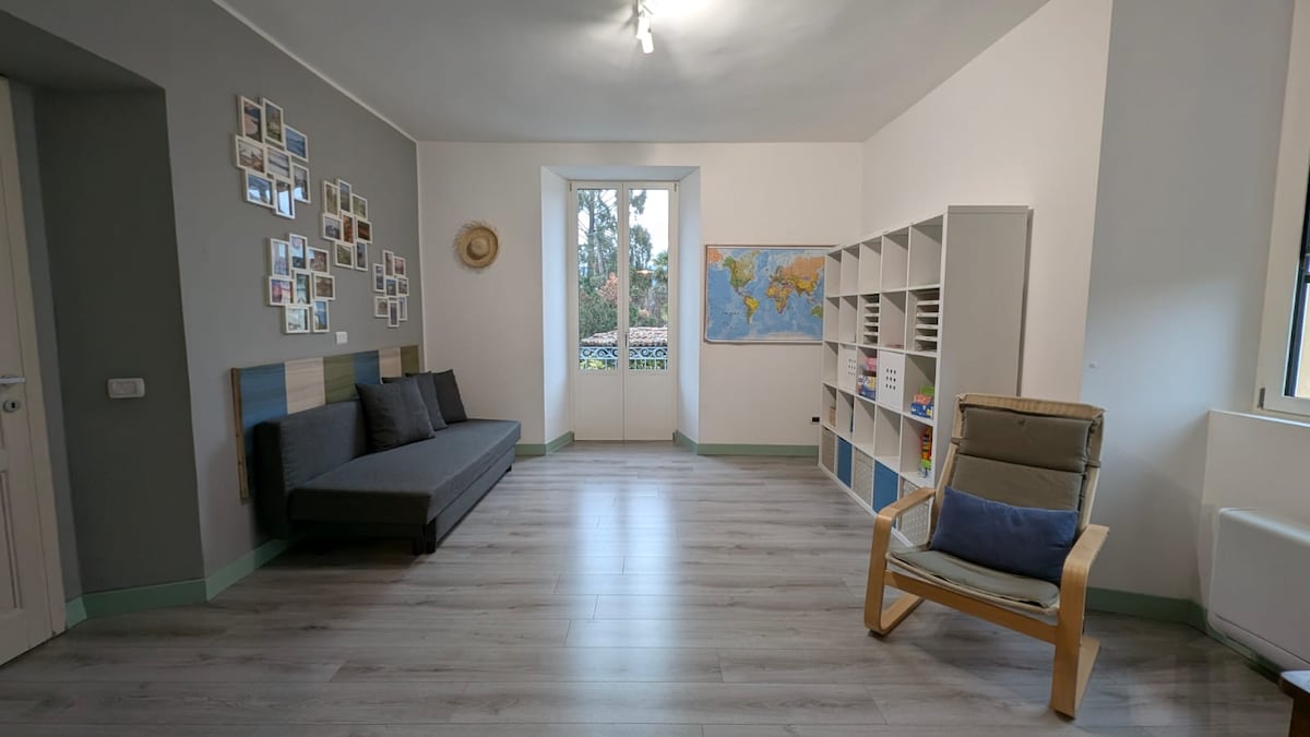 A spacious living area features a foldable sofa and a comfortable armchair positioned near a large window allowing natural light to enter. A bookshelf and a world map are displayed on the walls, enhancing the room’s inviting and airy atmosphere.
