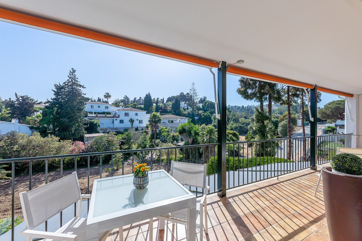 A spacious terrace is shown, furnished with a glass-top table and four white chairs. Greenery and residential buildings are visible in the background, providing a serene outdoor setting. The area is bathed in natural light, creating a welcoming atmosphere for relaxation.