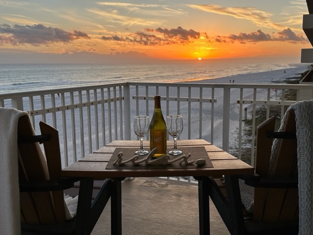 A balcony table is set for two, featuring a bottle of wine and two glasses, with a woven placemat beneath. A stunning sunset is visible over the ocean, casting warm hues across the sky and reflecting on the water. The beach stretches in the background.