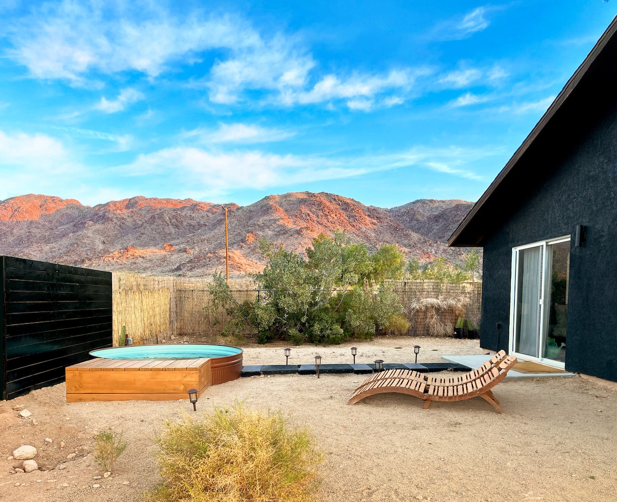 The outdoor space features a cowboy pool and a wooden lounge chair on a sandy area, surrounded by desert vegetation. The backdrop includes the colorful mountain range under a bright blue sky, creating a serene atmosphere for relaxation.