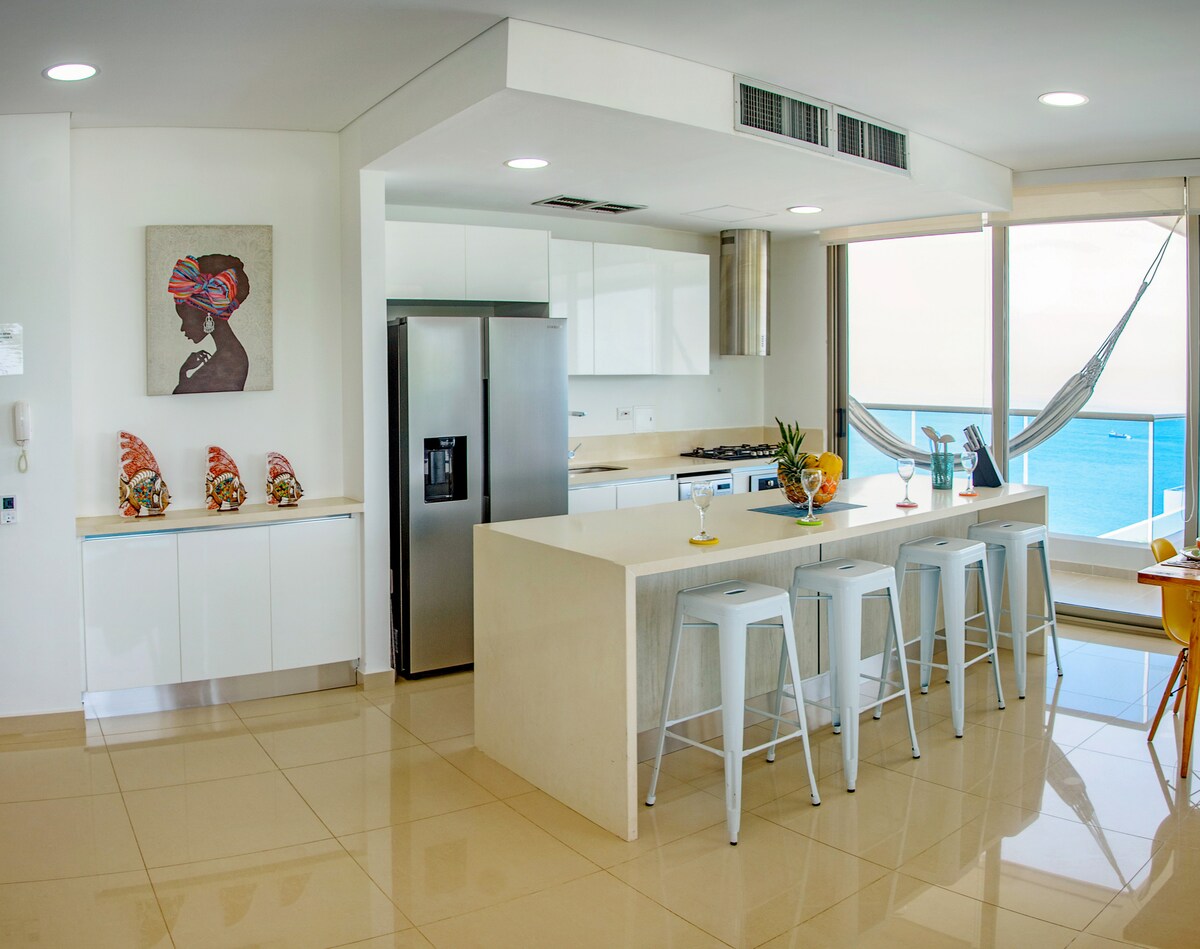 A modern kitchen is showcased, featuring white cabinetry and a large island with four stools. Fresh fruits in a bowl are placed on the counter. A stylish wall art piece is visible, along with a view of the sea through spacious windows. Natural light fills the room.