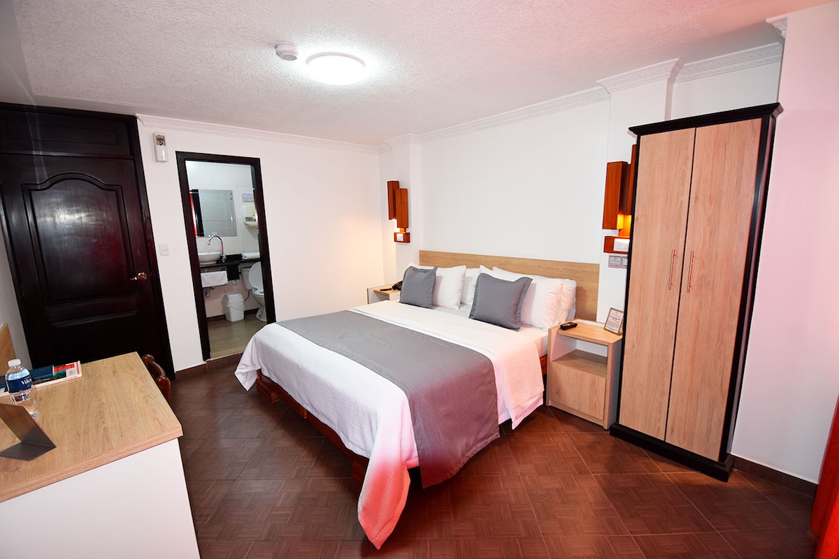 A spacious room features a large bed with neutral-toned bedding. A wooden nightstand and a wardrobe add functional style. Warm lighting is provided by wall sconces, and an en-suite bathroom is visible through the open door.