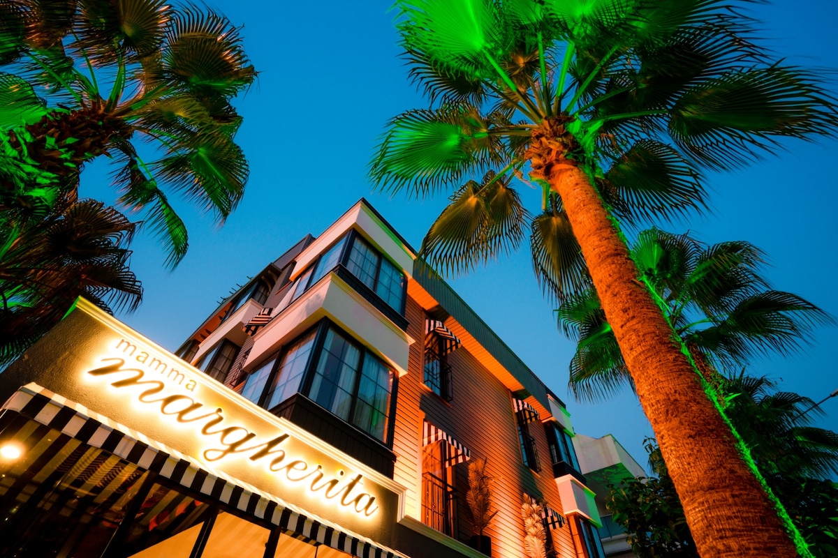 The exterior of a multi-story building is showcased, featuring a prominent sign reading 'mamma margherita.' Lush palm trees frame the image, illuminated by green lighting. The ambiance suggested by the vibrant colors hints at a lively atmosphere, welcoming guests with a tropical feel.