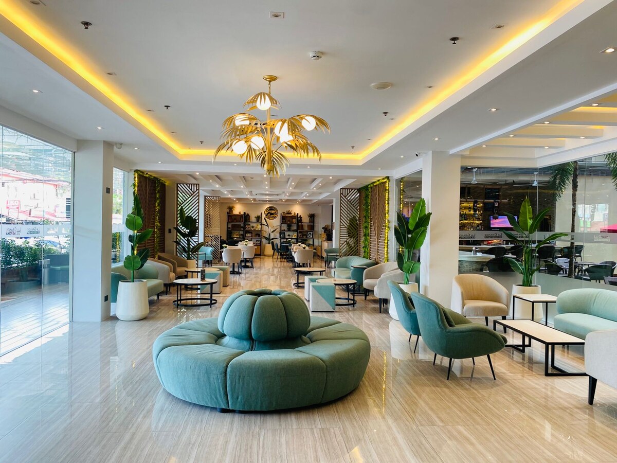 An inviting lobby area features a plush circular seating arrangement at the center, surrounded by stylish chairs and small tables. Soft lighting highlights the modern design elements, while lush indoor plants add touches of greenery to the space.