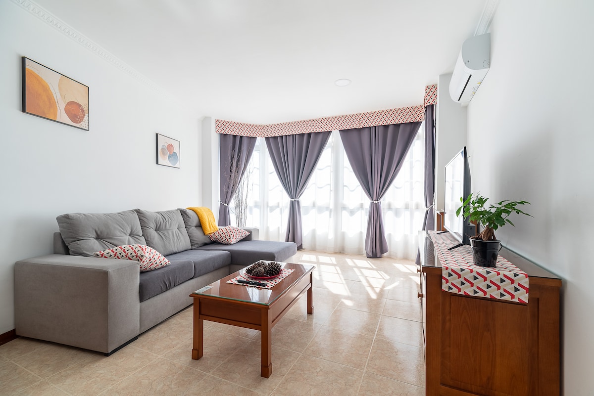 A bright living area features a large sectional sofa adorned with patterned cushions. A wooden coffee table occupies the center, complemented by a decorative arrangement. Light filters through the sheer curtains, highlighting the simplicity of the space, which includes a wall-mounted television and a small potted plant.