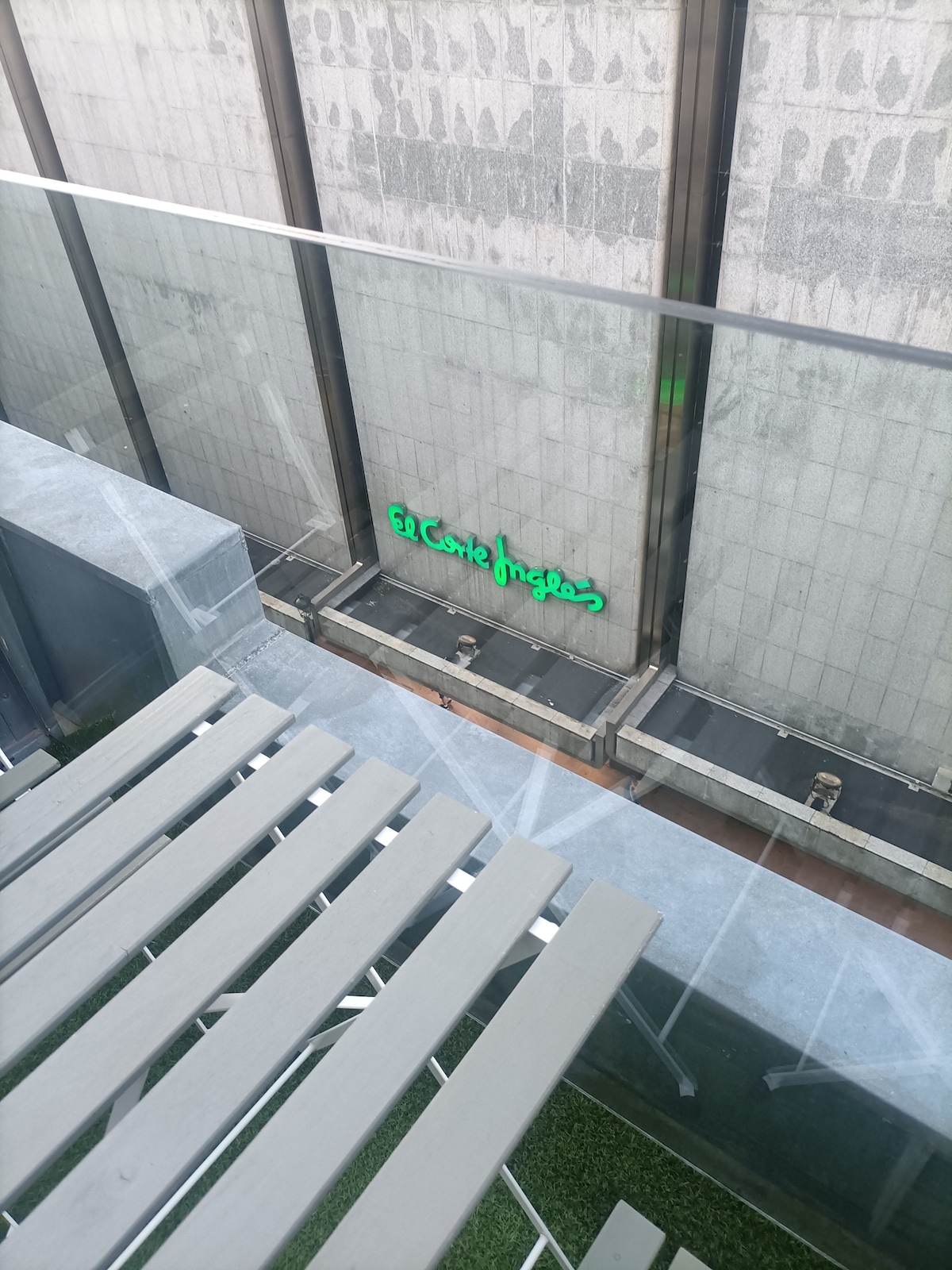 A glass balcony railing is visible, providing a view of a shopping center sign. The ground below features grey concrete and a touch of greenery. The neon green sign of 'El Corte Inglés' stands out against the building's facade, indicating the proximity to urban amenities.