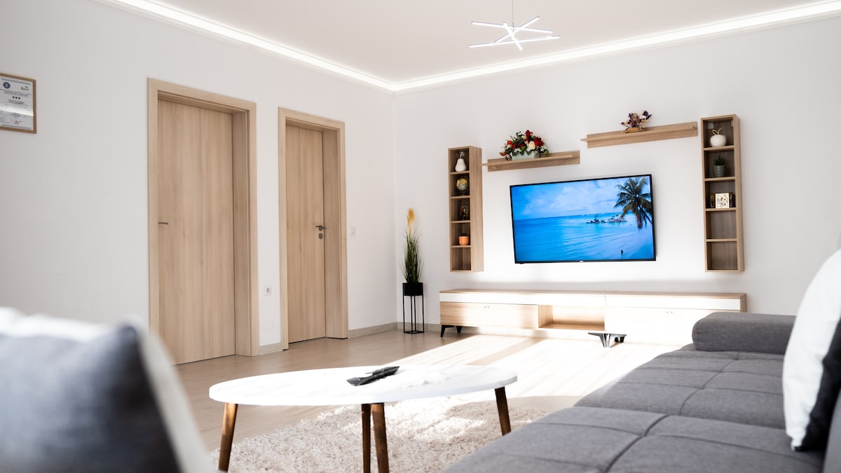 A modern living area is presented, featuring a large wall-mounted smart TV and light wooden entertainment unit. Decorative elements are arranged on the shelves, while natural light illuminates the space through neutral-colored walls. A cozy sofa and coffee table create a welcoming seating area.