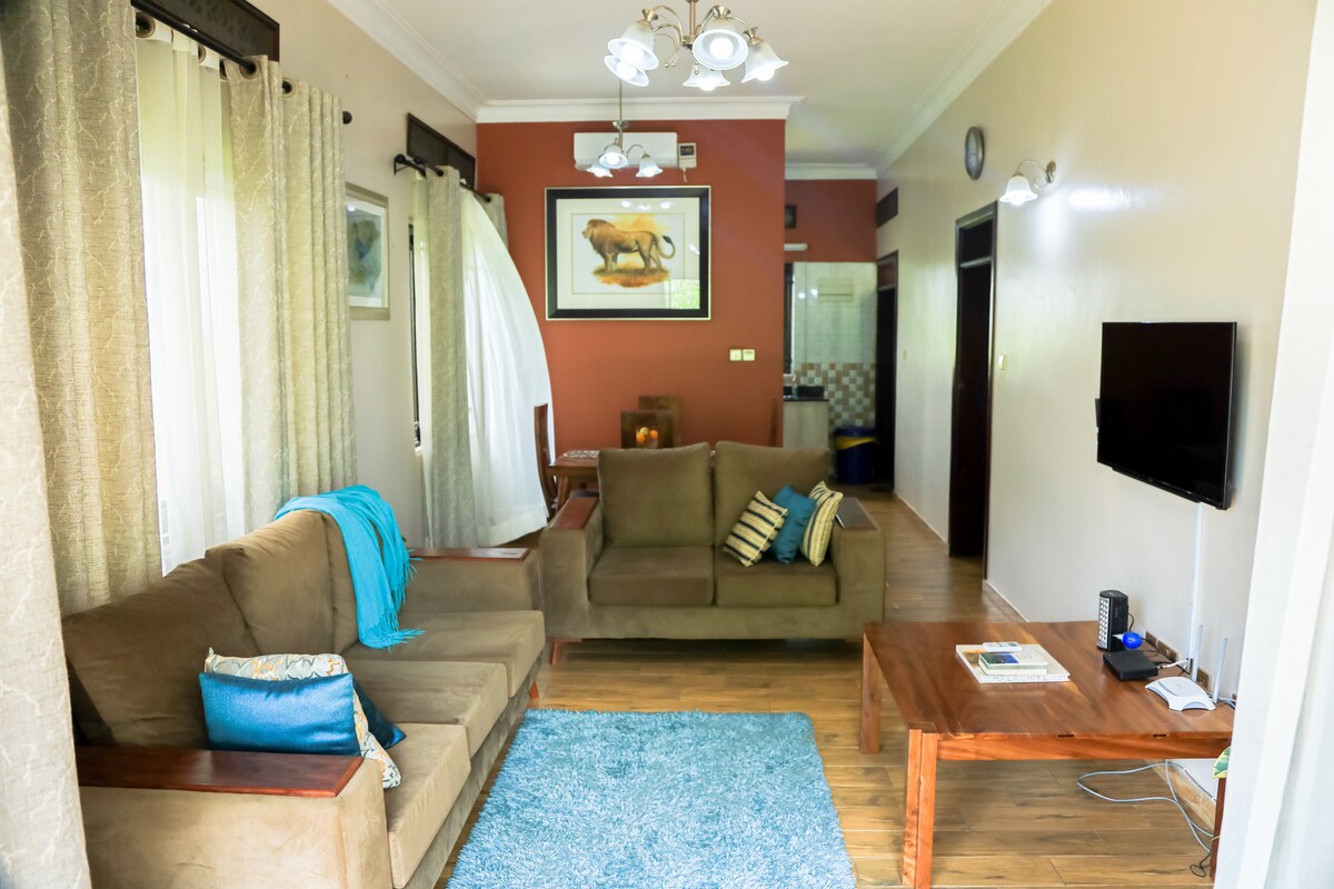 A spacious living area features two soft, neutral-colored sofas accompanied by decorative cushions. A coffee table rests on a soft blue rug, while natural light filters in through sheer curtains. A small wall-mounted television and artwork add character to the room.