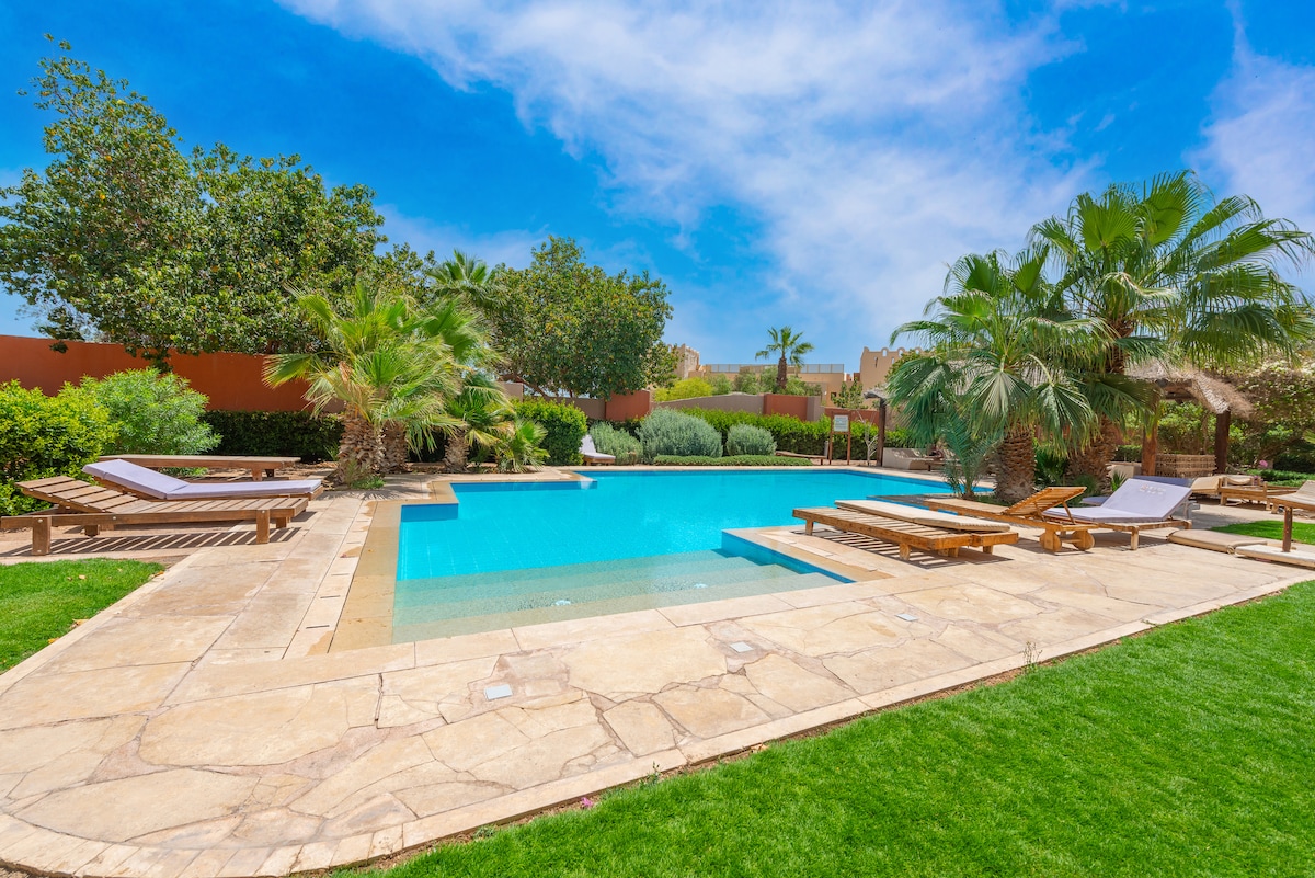 A serene communal pool area is surrounded by lush greenery and palm trees. Sun loungers are positioned alongside the pool, offering ample space for relaxation. The pool reflects the blue sky, creating a calming atmosphere for guests to enjoy.