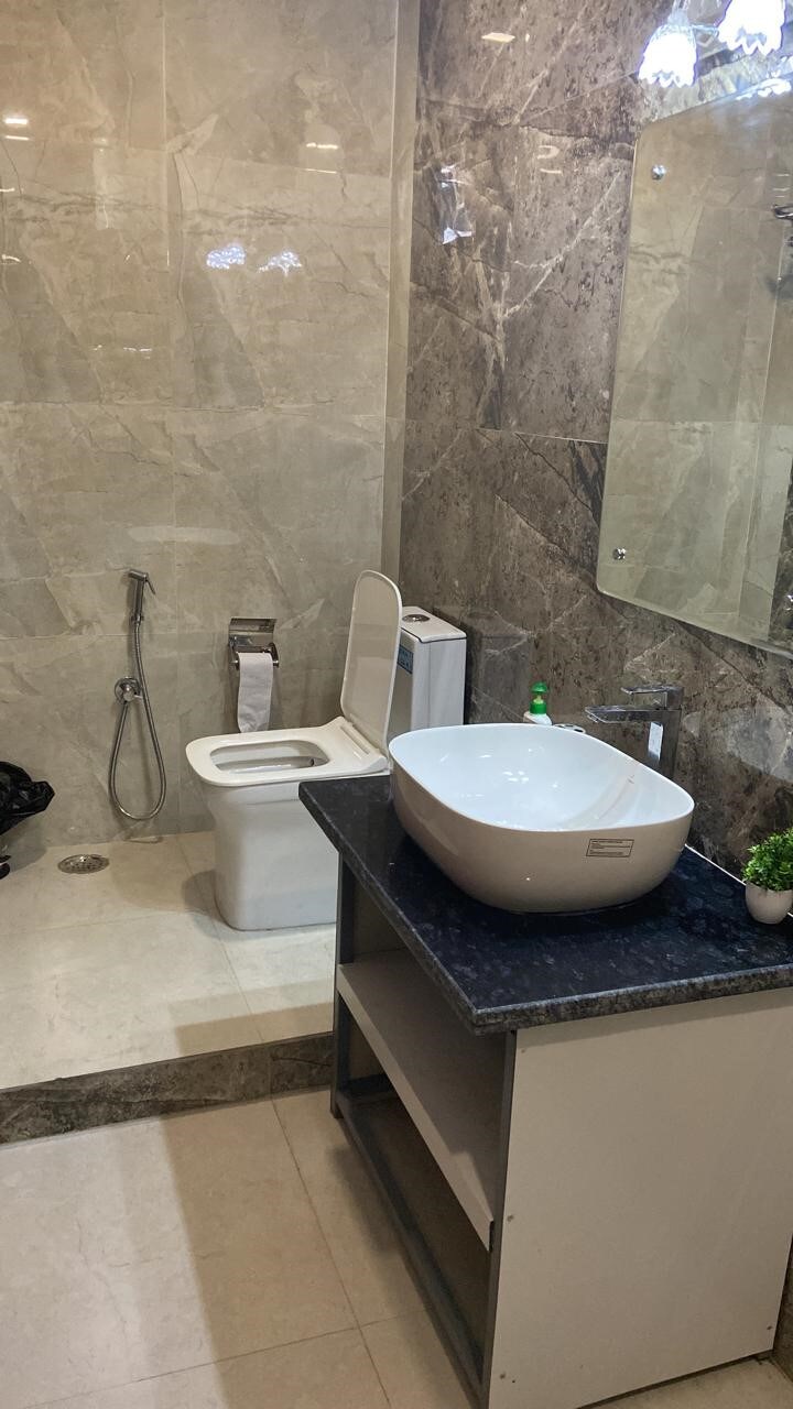 A modern bathroom is configured with sleek marble walls and a spacious shower area. A white vessel sink rests atop a dark countertop, complemented by a small potted plant. A toilet is positioned next to the sink, with an elegant mirror mounted above.