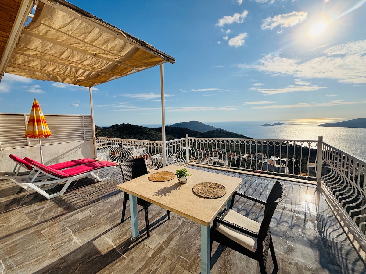 An outdoor terrace features a dining table set for four, complemented by a sun umbrella and lounge chairs. A scenic view of the coastline and water reflects the sunlight, providing a peaceful setting. The surrounding area includes mountains and distant islands.