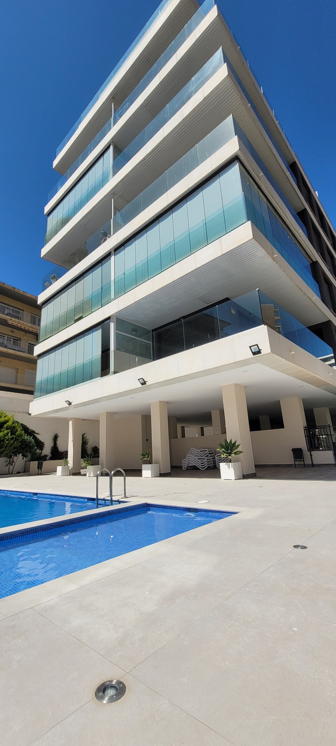 A modern building features several glass balconies, providing ample natural light and a view of the surrounding area. A well-maintained swimming pool is situated in front of the structure, surrounded by a tiled deck and potted plants.