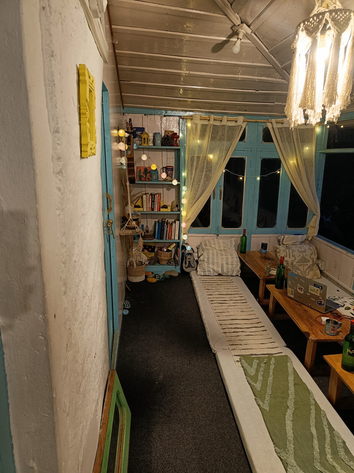 A cozy common area is highlighted by light-colored walls and soft, ambient lighting. Shelves filled with books line the wall, while small wooden tables are positioned beside cushioned seating. Decorative string lights enhance the atmosphere, creating a welcoming space for relaxation.