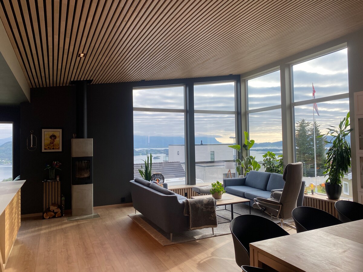 A modern living area is highlighted by large windows that offer expansive views of the surrounding landscape. The space features sleek furniture, including a comfortable couch and chairs, along with a cozy fireplace. Natural light enhances the inviting atmosphere throughout the room.