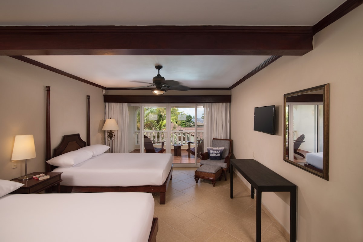 A room with two spacious beds is complemented by a ceiling fan and a cozy seating area. Natural light filters in through a glass door leading to a private balcony, while a flat-screen TV and a small desk provide functional amenities.