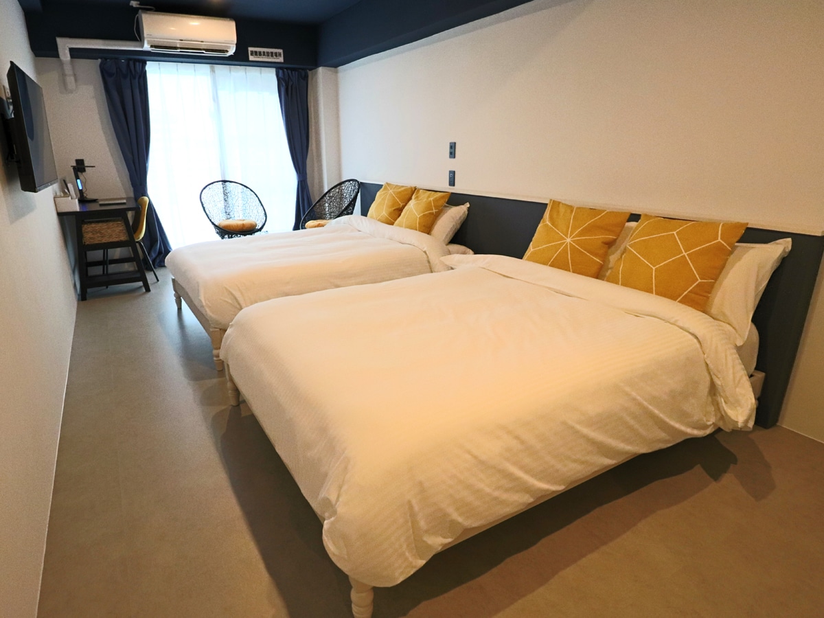 A room featuring two spacious beds, each adorned with yellow accent pillows. Natural light fills the space through a large window with sheer curtains. A small table and two stylish chairs are positioned nearby, adding to the room's inviting layout.