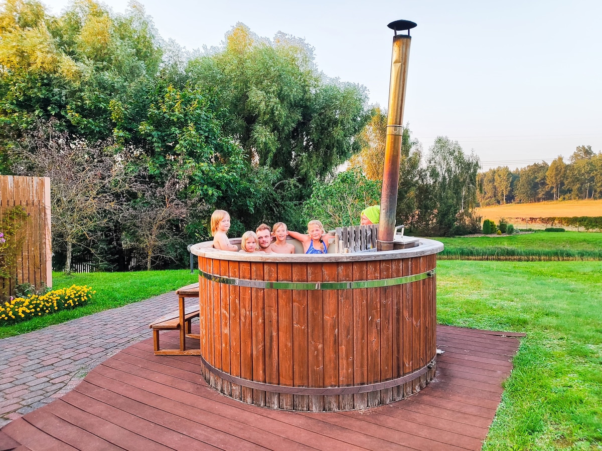 A wooden hot tub is surrounded by a grassy area and trees, inviting guests for relaxation. The tub features a chimney for warmth, with several individuals enjoying the experience. A paved path leads to the tub, enhancing accessibility.
