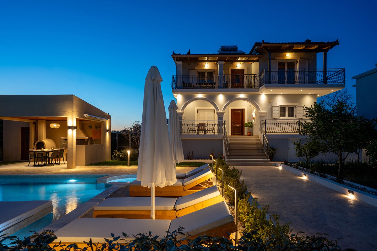 A two-story villa is softly illuminated against a twilight sky. The pool area is highlighted with lounge chairs and a protective umbrella. Pathways lined with lights lead to an outdoor dining area, while the facade displays a welcoming entrance with arched balconies.