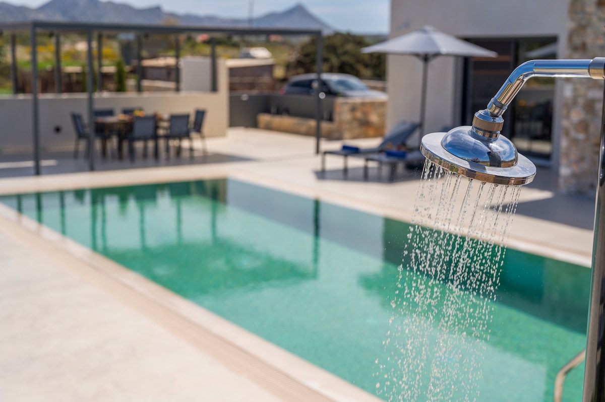An outdoor shower is positioned beside a spacious, clear pool. Lounge chairs and a dining area with a shaded table are visible in the background, complemented by lush greenery and a tranquil atmosphere.