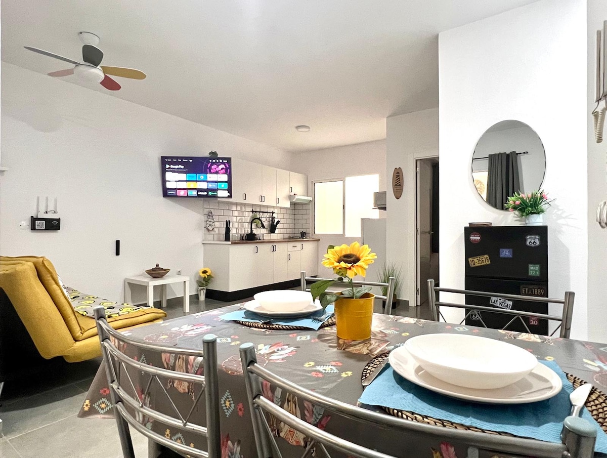 A modern living space is seen with a dining table set for a meal, featuring floral decor. The kitchen is equipped with sleek cabinetry and modern appliances. A smart TV is mounted on the wall, while a comfortable seating area includes a vibrant yellow armchair.