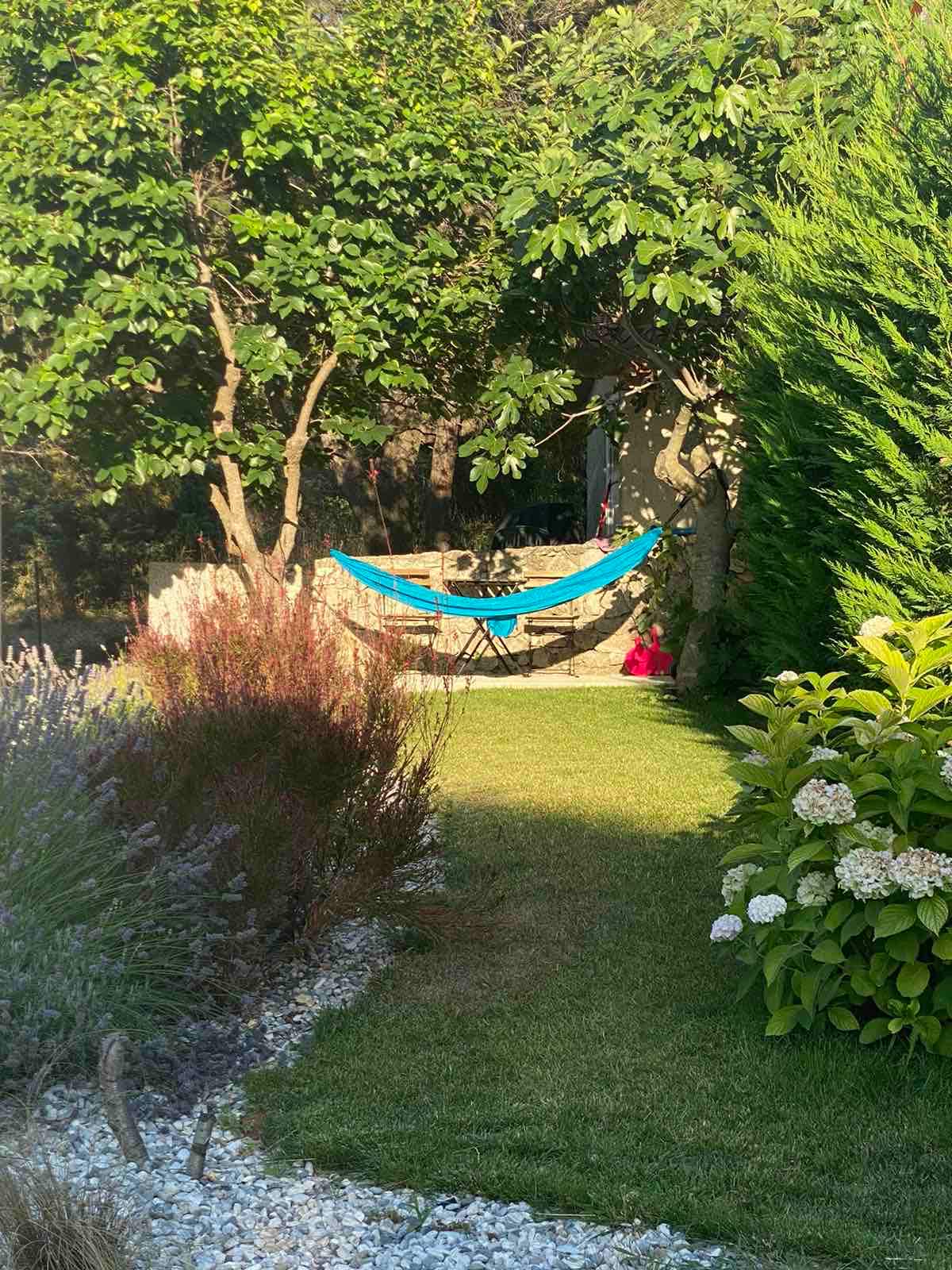 A serene garden space is showcased, featuring a vibrant blue hammock suspended between two trees. Lush green grass and various Mediterranean plants are present, including flowering shrubs and lavender. Pebbles line part of the pathway, offering a natural look to the outdoor area.