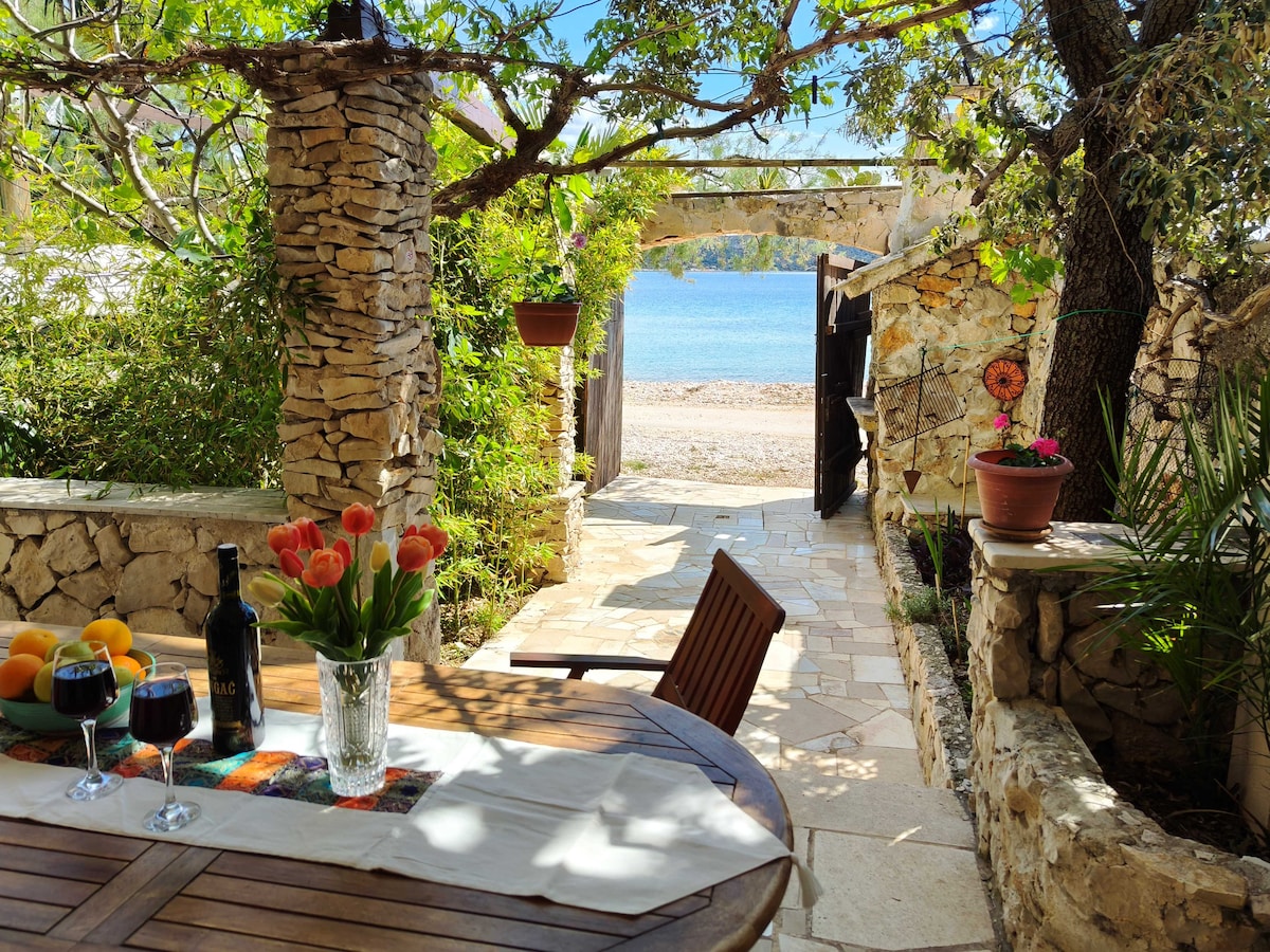 A serene outdoor dining area is surrounded by stone walls, featuring a wooden table set with two glasses and a bottle. Bouquets of tulips and fresh oranges are on display. A pathway leads to the crystal-clear waterfront, framed by lush greenery and shaded trees.