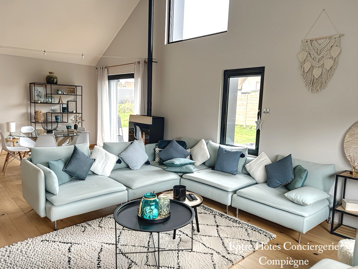 A spacious living area features a light blue sectional sofa adorned with various decorative cushions. A round black and white patterned rug anchors the space, complemented by a glass coffee table. Behind, a minimalist shelving unit displays decorative items, and large windows allow natural light to fill the room.