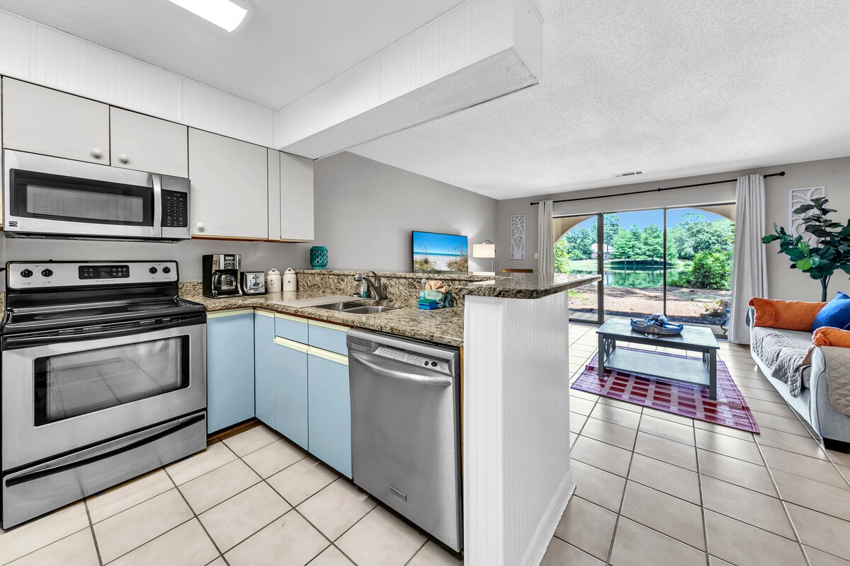 The kitchen area is equipped with modern appliances, including a stainless-steel microwave, oven, and dishwasher. Light cabinetry contrasts with a granite countertop. A living area with a sofa and a decorative table showcases a view of the outdoor patio through sliding glass doors.