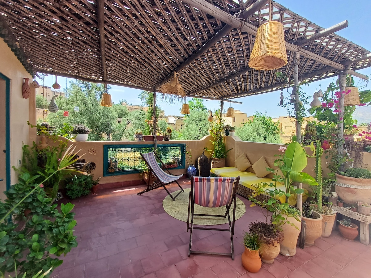 A serene terrace is adorned with lush greenery and potted plants, featuring comfortable seating areas. A striped chair and a sun lounger invite relaxation under a pergola, which is decorated with hanging lanterns. The space offers views of ancient olive trees and the surrounding landscape.
