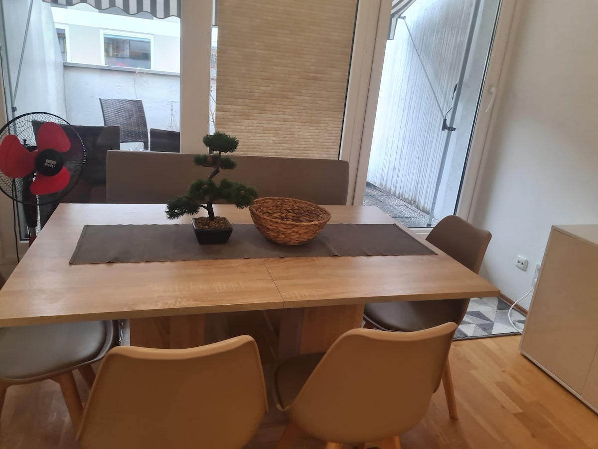 A modern dining area is featured with a light wood table set for six. A decorative basket and a small plant adorn the center, enhancing the space. Large windows bring in natural light, and a fan is positioned nearby for comfort.
