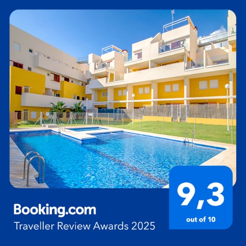 An outdoor swimming pool is framed by a well-maintained grassy area, surrounded by the colorful yellow and white buildings of the complex. Sun loungers are arranged alongside the pool, inviting relaxation in a tranquil environment.