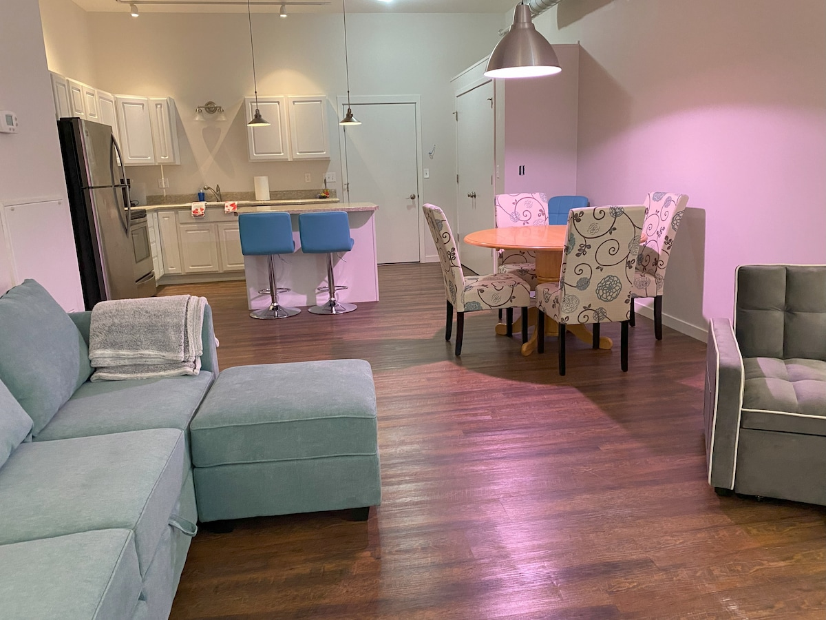 An open living area is displayed, featuring a sectional sofa in soft gray. A round dining table with decorative chairs is positioned near the kitchen, which includes modern appliances and a bar counter with stools. Warm lighting enhances the wood flooring throughout the space.