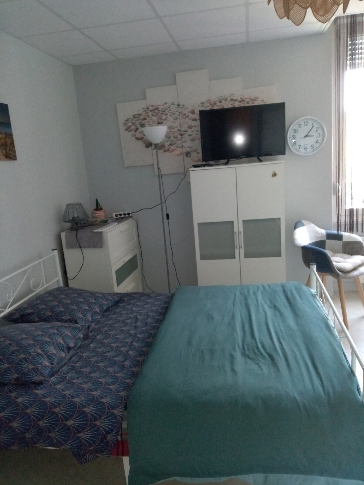 The studio bedroom features a spacious bed with patterned bedding, complemented by a modern floor lamp nearby. A flat-screen TV is mounted on the wall above a stylish white cabinet. A simple clock and a comfortable chair are positioned in the room, allowing for relaxation.