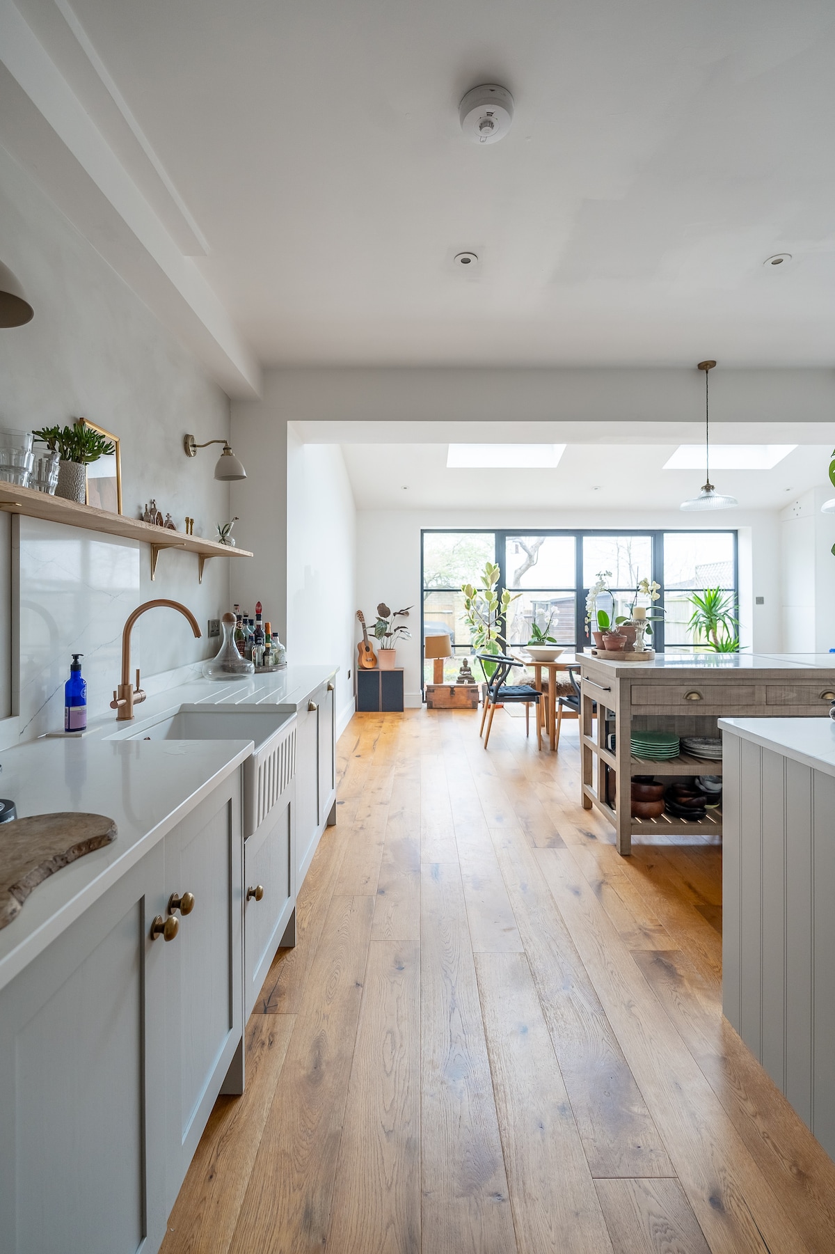 The open-plan kitchen and dining area is illuminated with natural light from skylights above. Elegant cabinetry lines the walls, complemented by modern fixtures. A dining table is positioned at the far end, surrounded by greenery, creating a welcoming atmosphere for cooking and gathering.