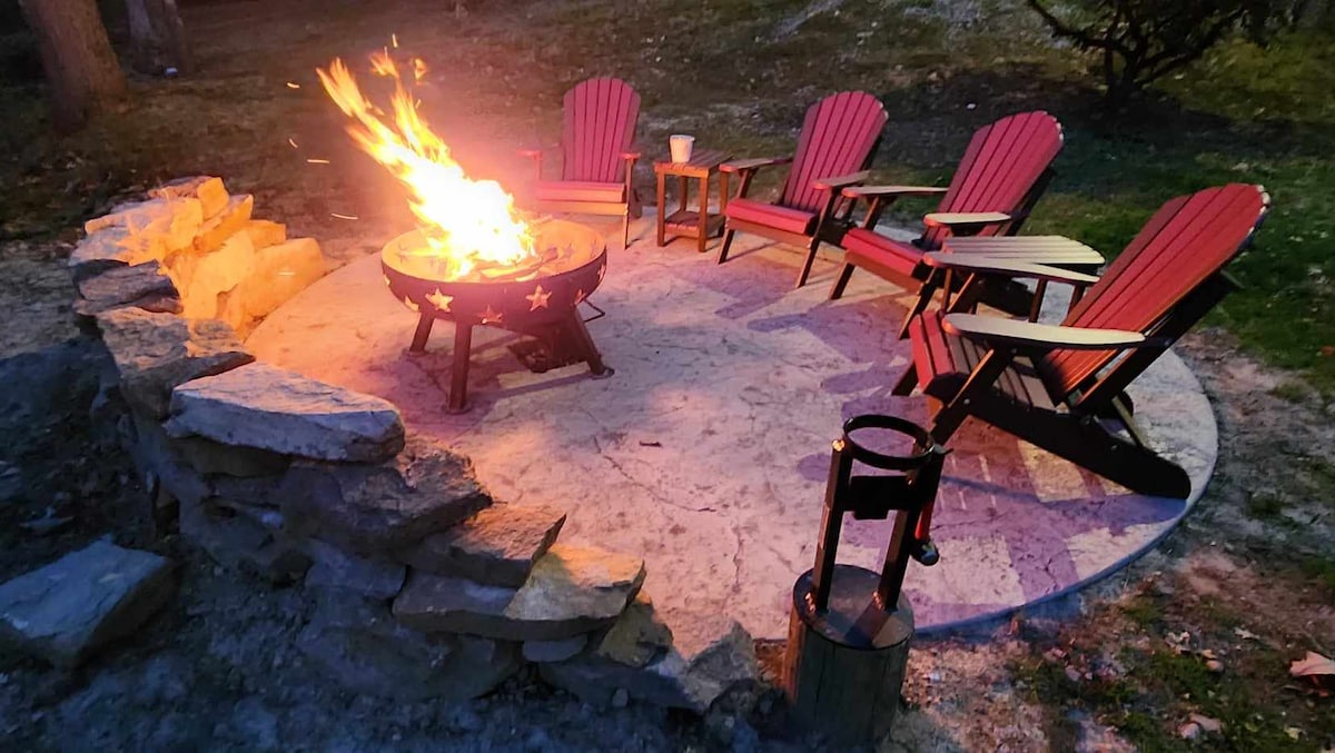 A circular stone firepit area is surrounded by five red Adirondack chairs. A fire burns brightly in the center, casting a warm glow. A small table can be seen beside one of the chairs, enhancing the cozy gathering space.