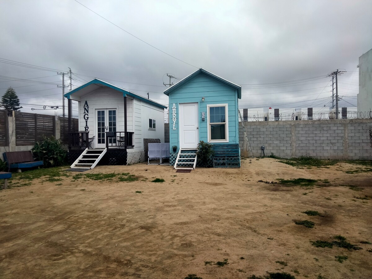 Two charming tiny cottages are depicted, each featuring distinctive color schemes. The one on the left is painted white with black accents, while the blue cottage on the right includes a white door and porch. A grassy area with sandy ground is shown in front.