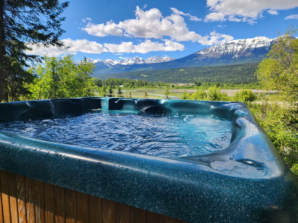 A hot tub is positioned outdoors, offering a scenic view of the surrounding mountains and valley. The water glistens under the sunlight, with lush greenery and distant snow-capped peaks visible in the background, creating a serene outdoor retreat.