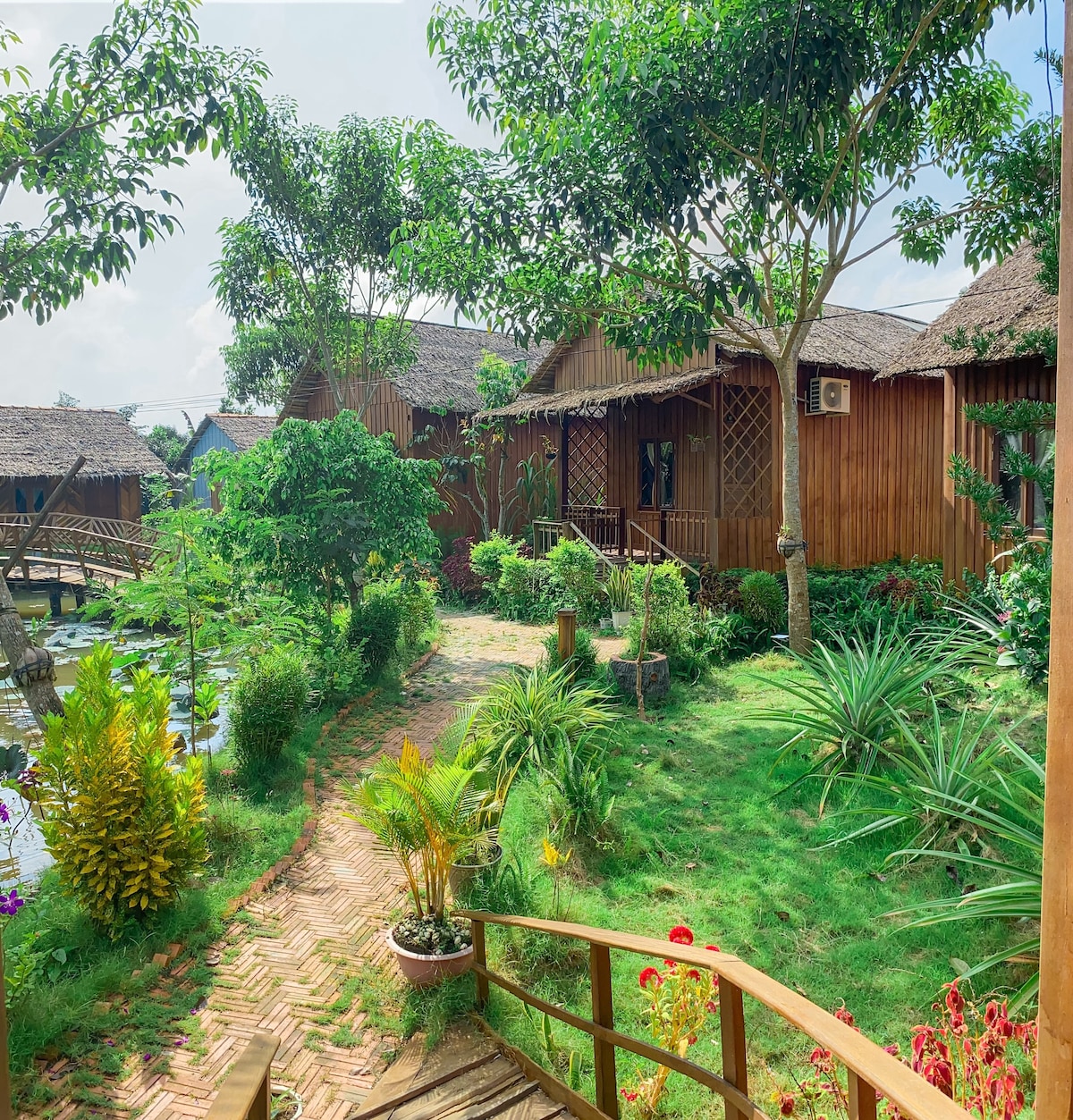 A serene landscape features a pathway meandering through lush greenery, leading to wooden cabin accommodations. Vibrant plants and trees surround the area, while thatched roofs peek through the foliage. The natural setting creates a peaceful atmosphere typical of the Mekong Delta countryside.
