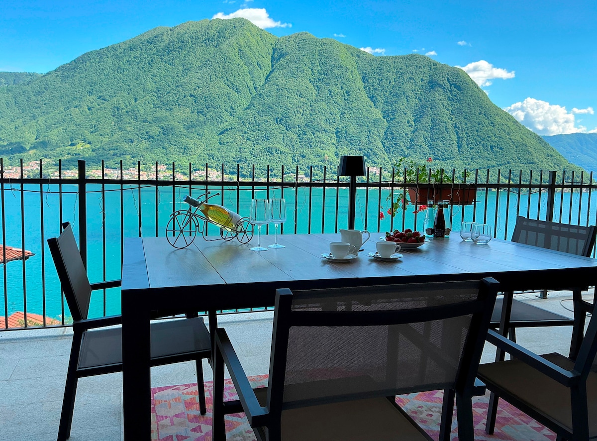 A spacious terrace is featured with a dining table set for four, overlooking the tranquil waters of Lake Como. Lush green mountains rise in the background, creating a serene view. Sun-loungers are positioned nearby, inviting relaxation in the open air.