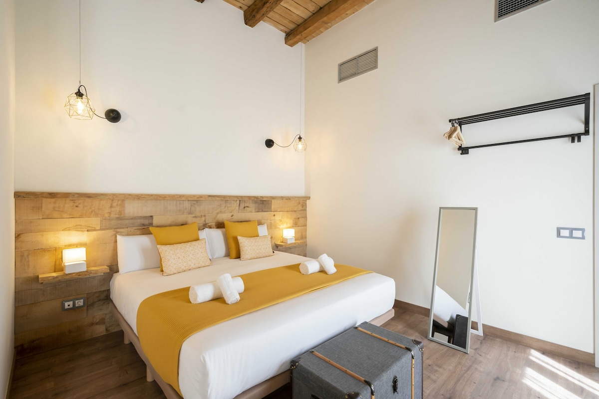 A spacious sleeping area features a large bed covered with a yellow bedspread and complemented by decorative pillows. Soft lighting is provided by wall sconces, while a full-length mirror stands nearby. Wood beams frame the ceiling, adding warmth to the modern decor.