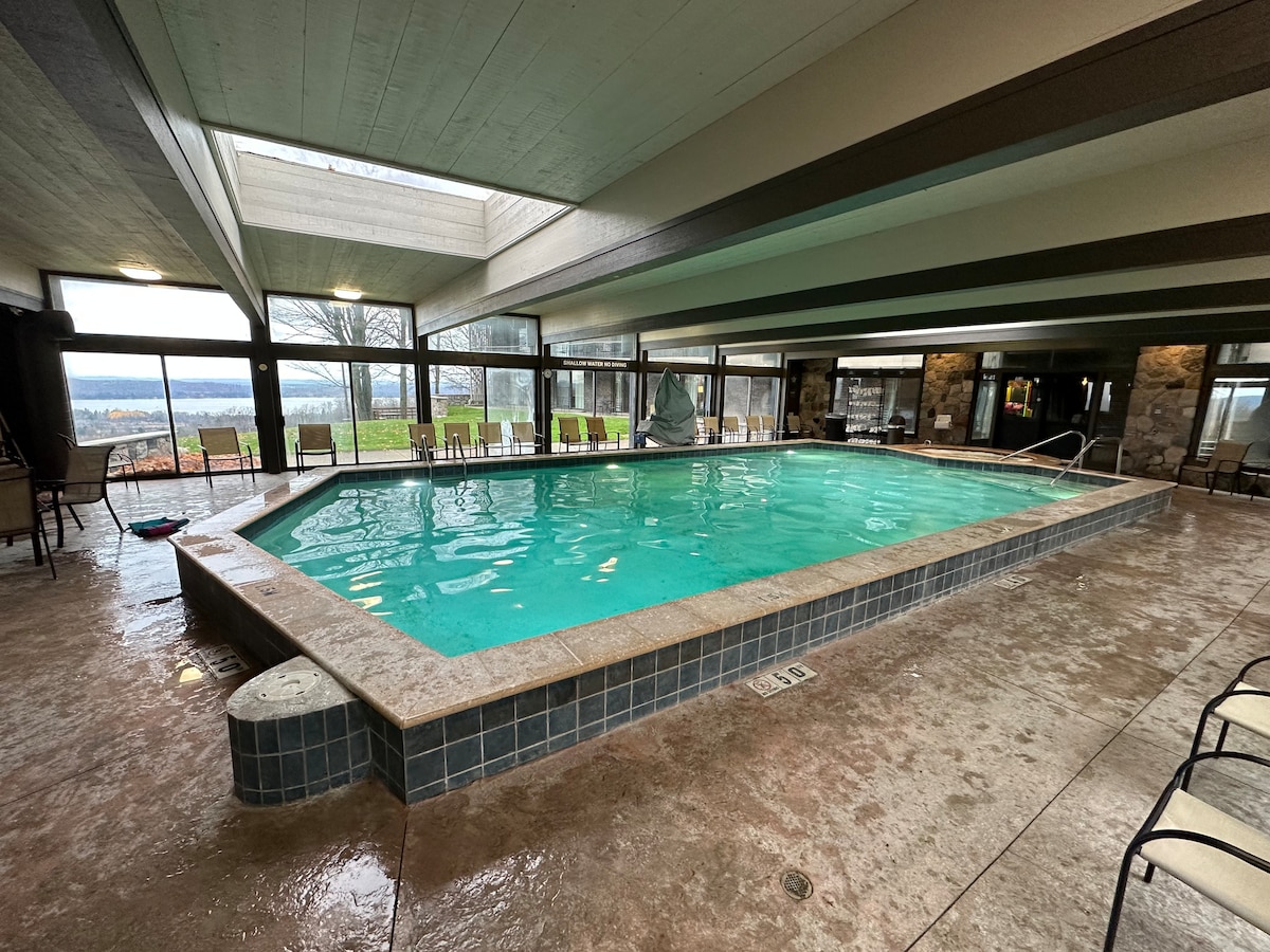 An indoor swimming pool is surrounded by large windows that provide expansive views of the outdoors. Natural light floods the area, accentuating the water's clear blue hue. Comfortable lounge chairs are positioned around the pool, inviting relaxation and leisure.