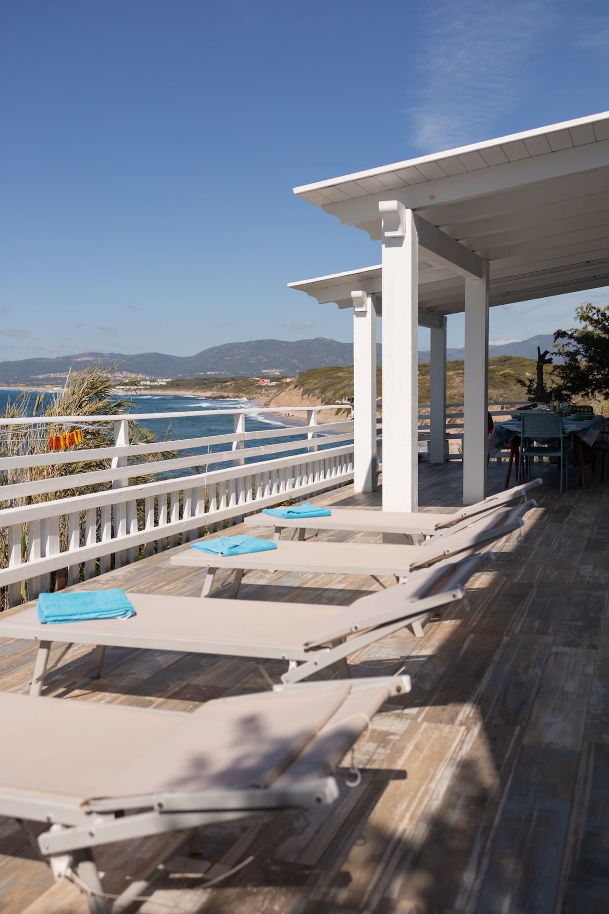 A spacious terrace features several lounge chairs with blue towels arranged on them. The area is complemented by white railings and overhead beams, offering scenic views of the coastline and mountains in the distance, bathed in natural sunlight.