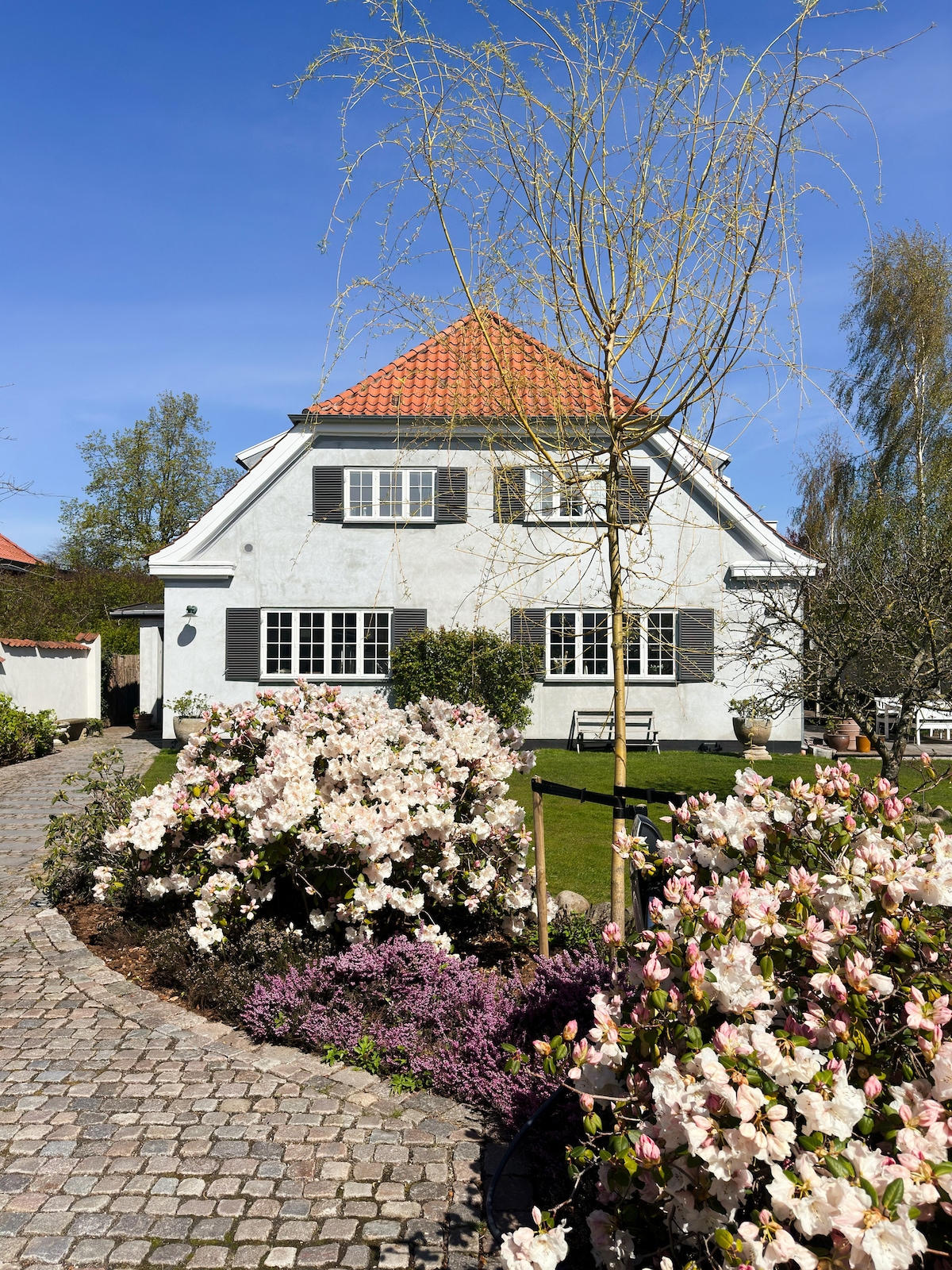 The exterior of the villa is shown, featuring a charming white facade with black shutters and a red-tiled roof. A cobblestone pathway leads through blooming gardens filled with pink and white flowers, while greenery surrounds the home, enhancing the serene atmosphere.