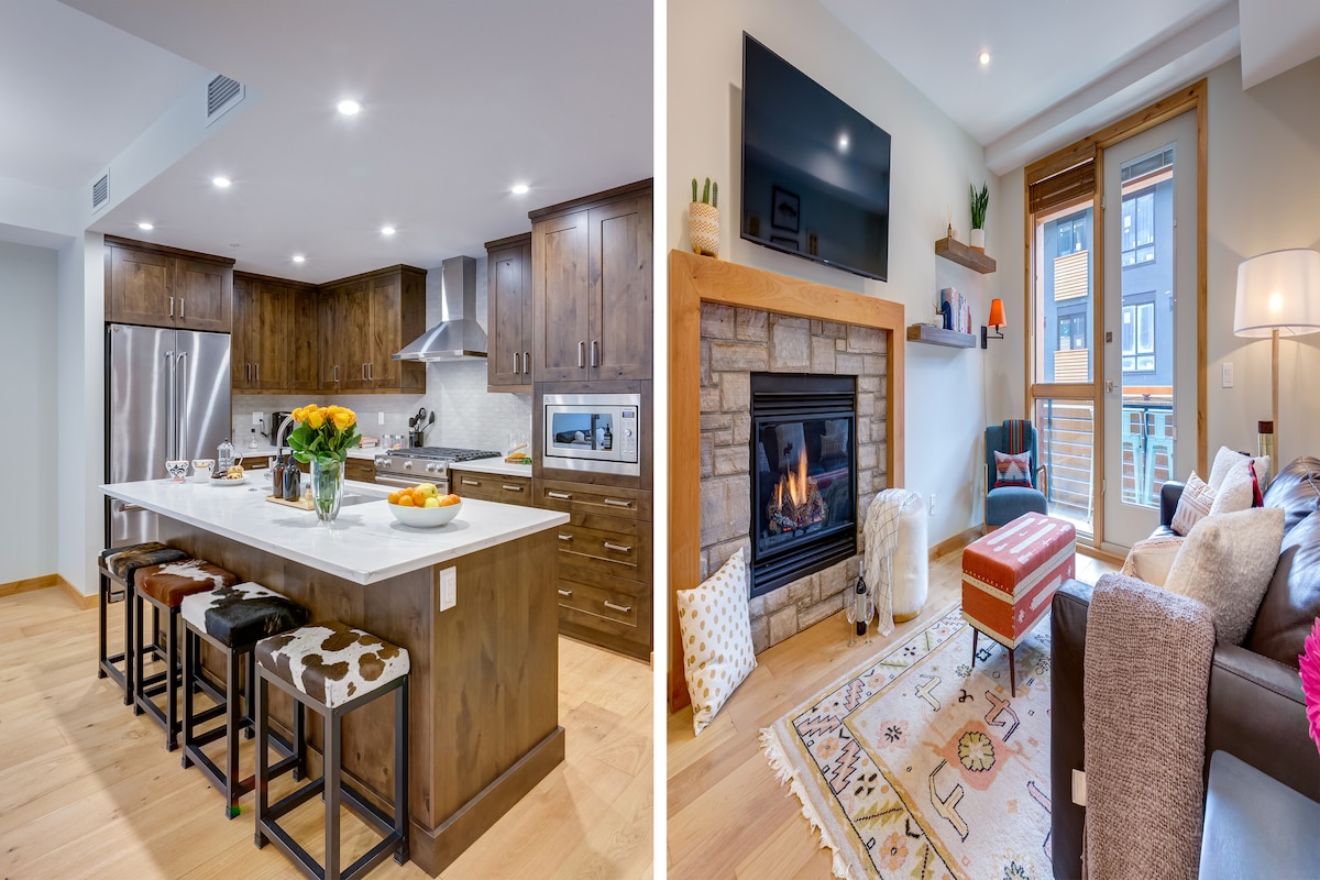 The kitchen features modern cabinetry with sleek wooden finishes and high-end appliances. A large island with seating is highlighted by natural light, creating an inviting space for cooking. Adjacent, the living area showcases a gas fireplace surrounded by stone, complemented by a cozy seating arrangement.